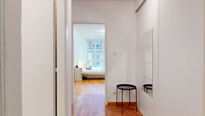 Ein enger Korridor mit Holzboden führt zu einem Schlafzimmer mit Bett, Nachttisch und Fenster. An der Wand stehen zwei schwarze Hocker.