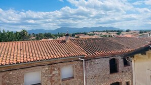 A view of terracotta-tiled rooftops, a stone building with a window, and distant mountains under a clear sky. - Appartement Neuf Bages Avec Balcon (Bages)