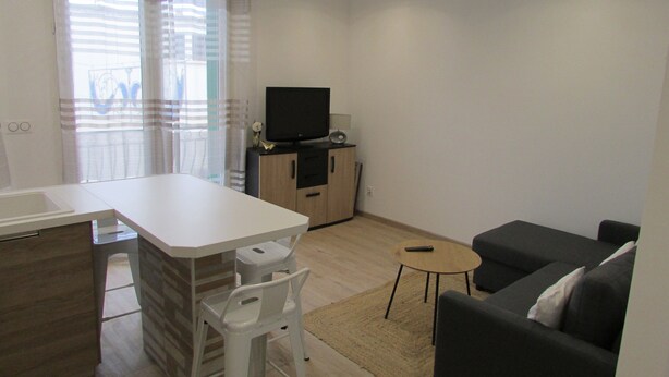 A modern living room with a sofa, a small round table, and a television on a cabinet. - Appartement Neuf Bages Avec Balcon (Bages)