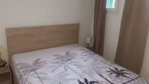 A single bed with a wooden headboard, a nightstand with a lamp and a vase of flowers, and a window with curtains. - Appartement Neuf Bages Avec Balcon (Bages)