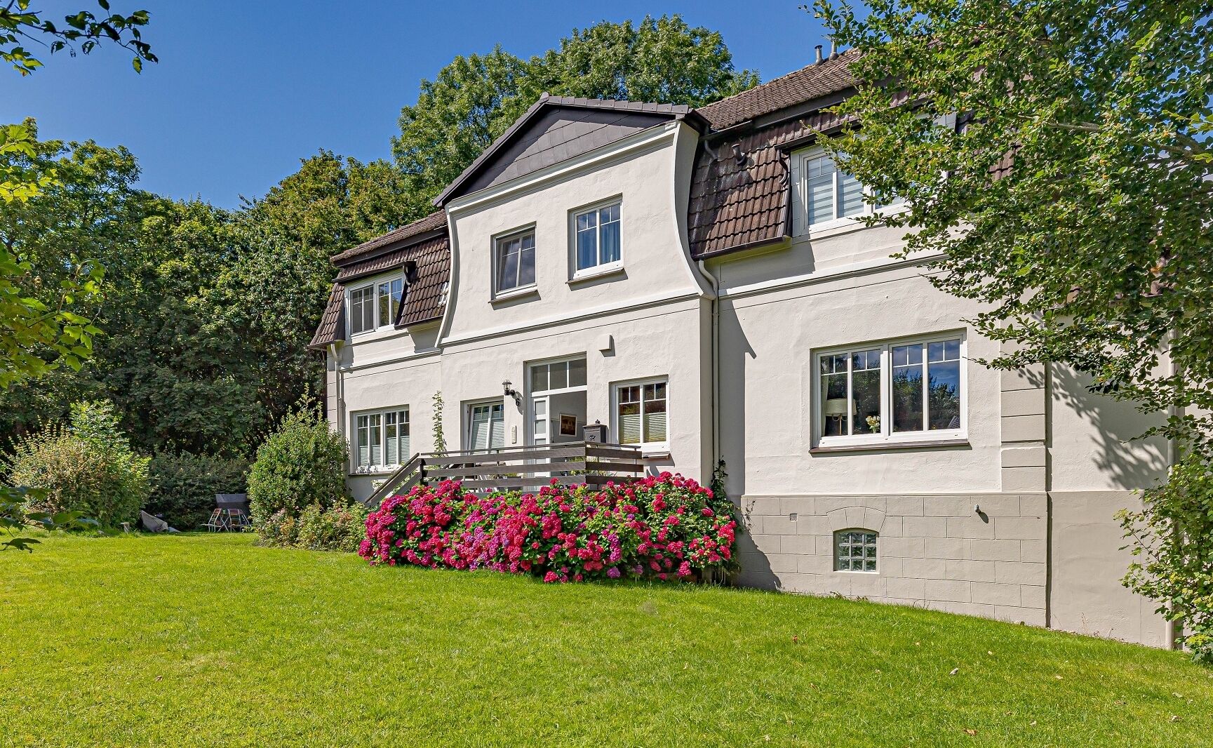 Une maison à deux étages avec un porche à l’avant, un petit jardin agrémenté de fleurs roses et une pelouse bien entretenue.