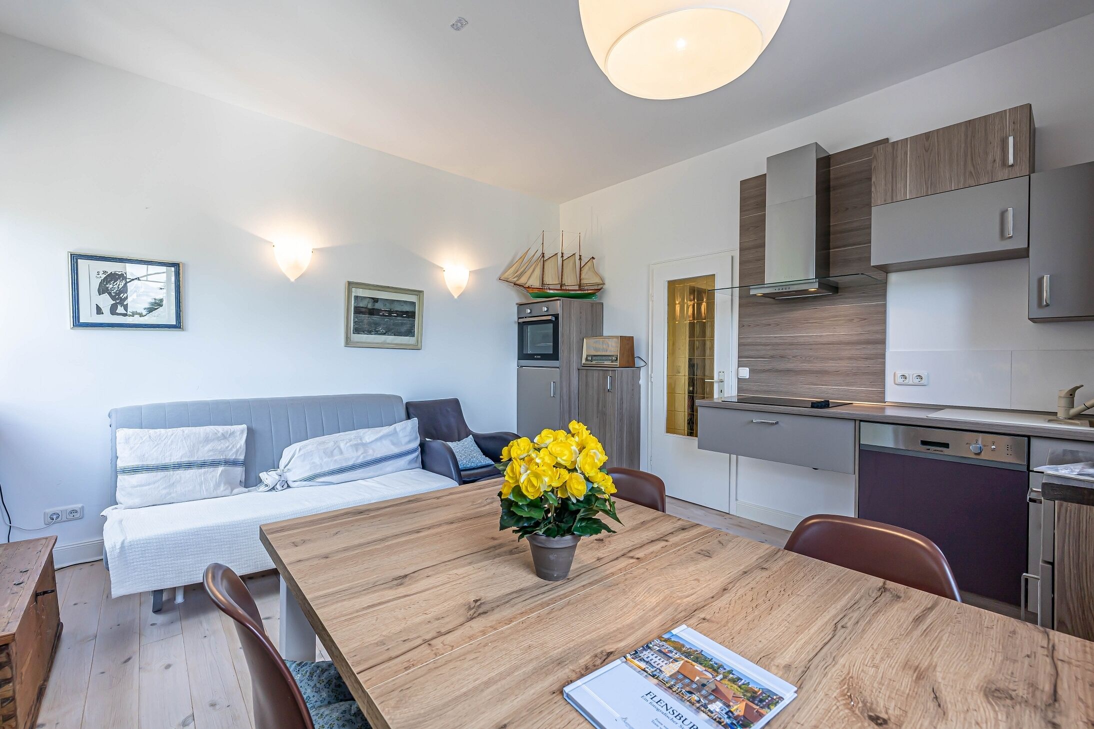 Un appartement moderne comprenant un coin repas, un canapé, une cuisine et une petite table à manger agrémentée d’un vase de fleurs.