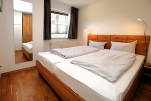 Un lit double avec du linge de lit blanc, une tête de lit en bois, une table de chevet avec une lampe, une fenêtre avec des rideaux et un miroir qui reflète la pièce.