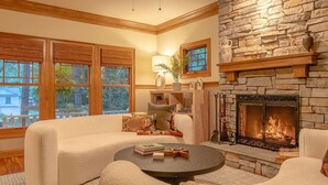 A cozy living room with a stone fireplace, a sofa, a coffee table, and a large window with blinds. - Your Michigan Cottage Awaits! (Douglas)