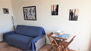 A living room with a blue sofa, a wooden table with a white vase, and four framed artworks on the wall. - Airy Studio with AC, Terrace, and Garage - Centrally Located in Agde (Cap d'Agde naturisme)