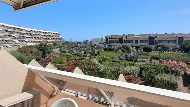 A balcony with a view of a landscaped garden, a modern building, and a clear blue sky. - Airy Studio with AC, Terrace, and Garage - Centrally Located in Agde (Cap d'Agde naturisme)