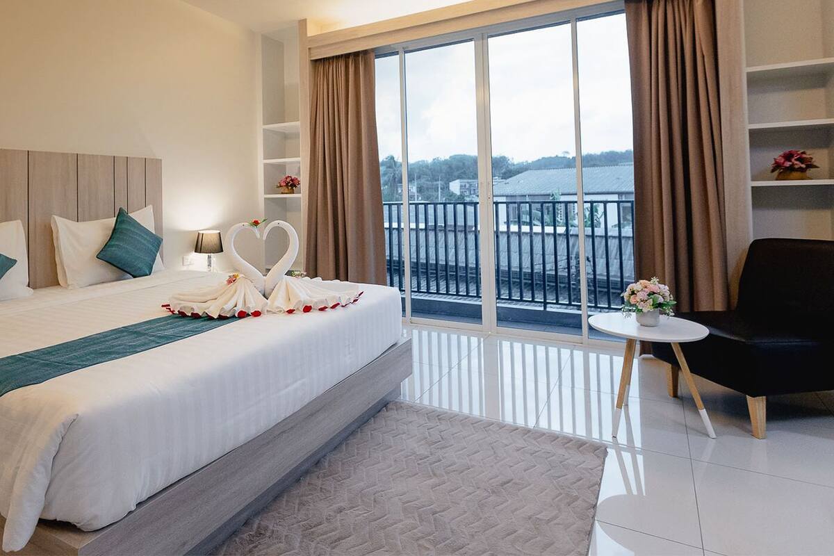 A hotel room with a large bed, a small round table, a black chair, and a balcony with a railing.
