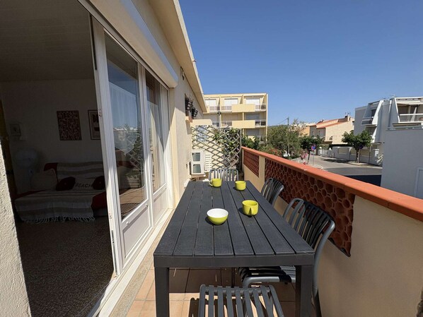 A balcony with a table and chairs, a sliding glass door, and a view of the street and other buildings.