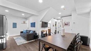 A modern kitchen with a built-in refrigerator, a dining area with a wooden table and chairs, a gray sofa with blue cushions, and a fireplace.