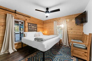 Quarto com painéis de madeira, cama, ventilador de teto, televisão montada e janela com cortinas.