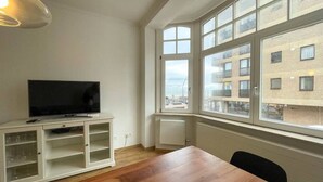Una stanza con un tavolo di legno, un armadietto bianco con vetri e una televisione a schermo piatto, oltre a una grande finestra con vista sugli edifici.