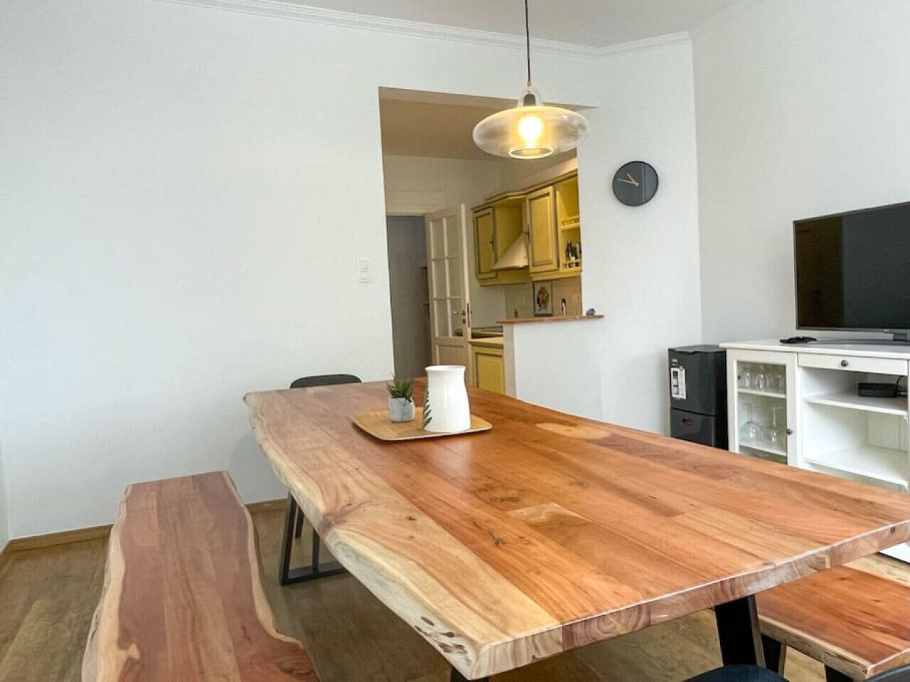 A dining area with a wooden table and bench, a hanging light fixture, and a kitchen in the background.