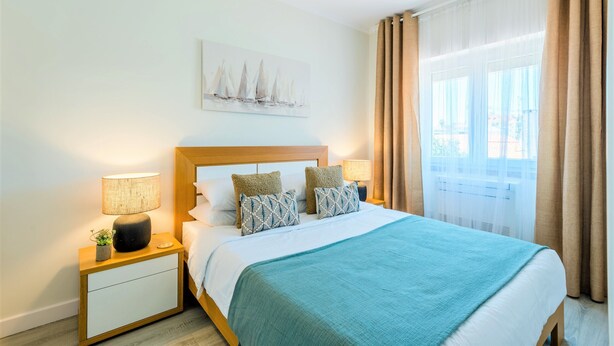 Une chambre d’hôtel avec un lit, deux tables de chevet, une commode, une lampe, une plante, un tableau et une fenêtre avec des rideaux.