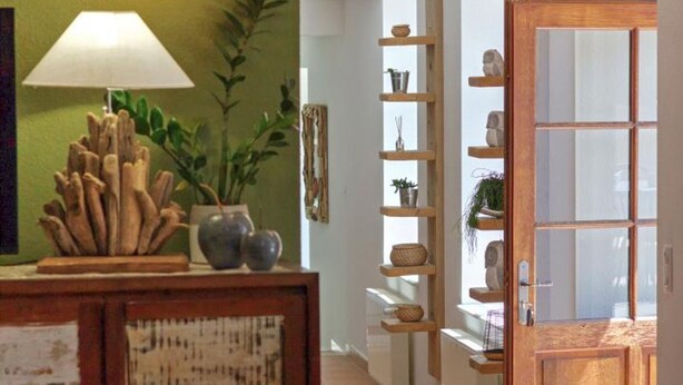 Um corredor com um armário de madeira exibindo objetos decorativos, uma planta em vaso e uma prateleira na parede com diversos itens.