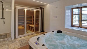 Uma casa de banho moderna com chuveiro de hidromassagem com parede de vidro, sauna de madeira, banheira grande com painel de controlo e janela que permite entrada de luz natural.