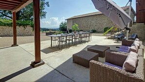 Um pátio externo com mesa e cadeiras, uma área de estar coberta com almofadas e um edifício de pedra ao fundo.