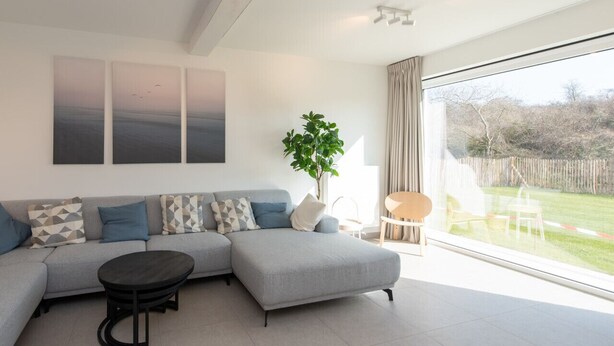 A modern living room with a grey sectional sofa, a round coffee table, and a large window with a view of a garden. - Villa Petit Mousse -Luxurious villa close to beach (Koksijde)
