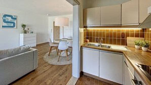Eine moderne Küche mit Holztischplatte und weißen Schränken, ein Essbereich mit rundem Tisch und Stühlen, und ein Wohnzimmer mit Sofa und Couchtisch.