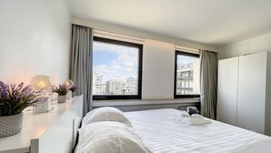 Ein modernes Hotelzimmer mit einem großen Bett, einem Schreibtisch mit Lampe und Blick auf die Stadt durch große Fenster.