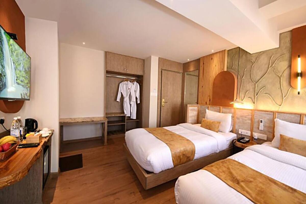 A hotel room with two beds, a TV mounted on the wall, a wooden desk with a basket of fruit, and a wardrobe with hanging robes.
