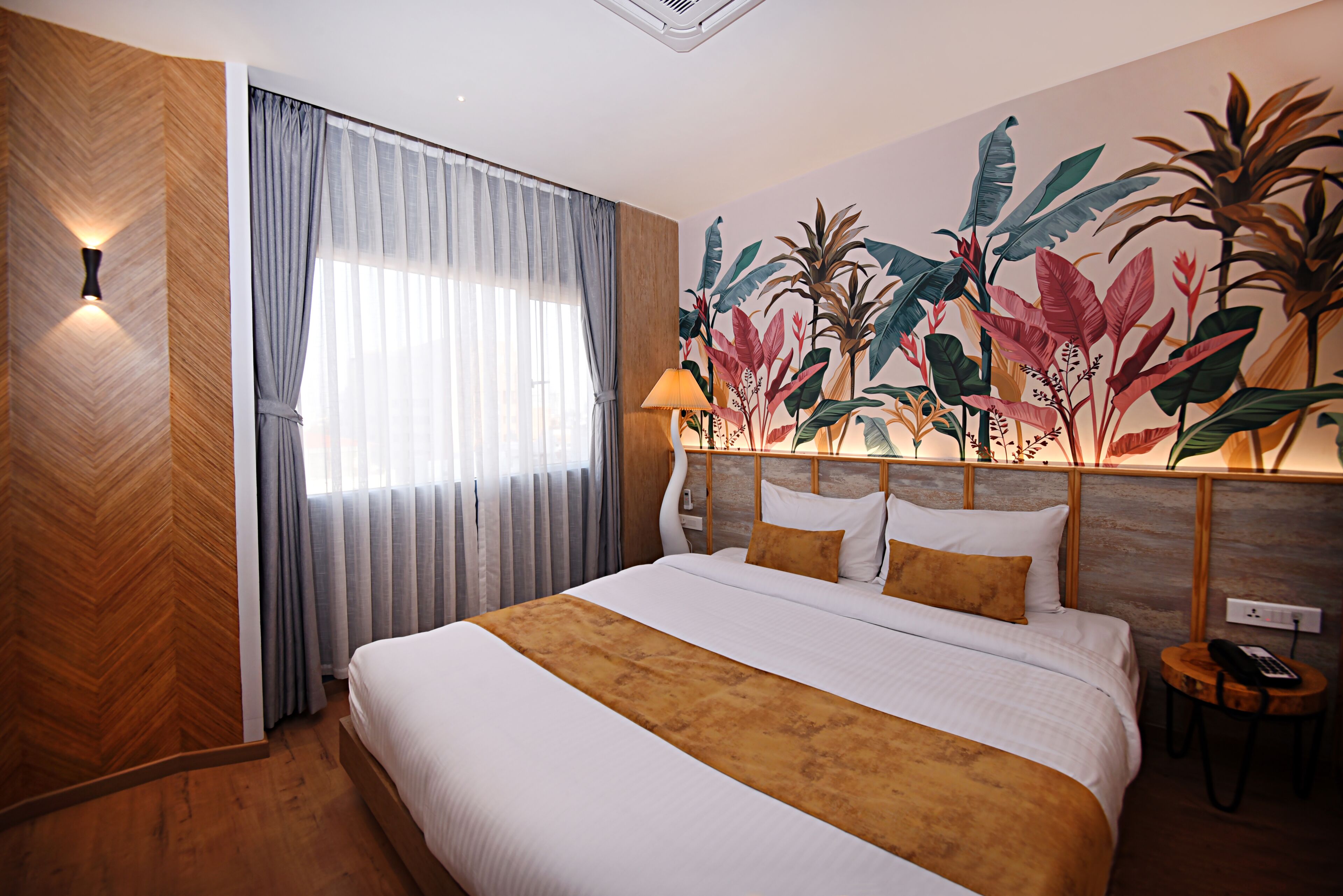 A hotel room with a large bed, a wooden headboard, a wall mural of tropical plants, a window with curtains, and a bedside table with a lamp.