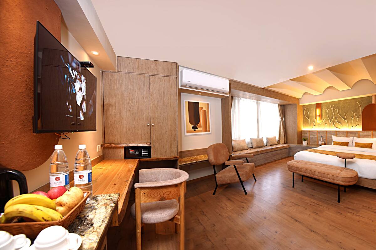 A hotel room with a wooden floor, a bed, a desk with a chair, a TV mounted on the wall, and a window with curtains.