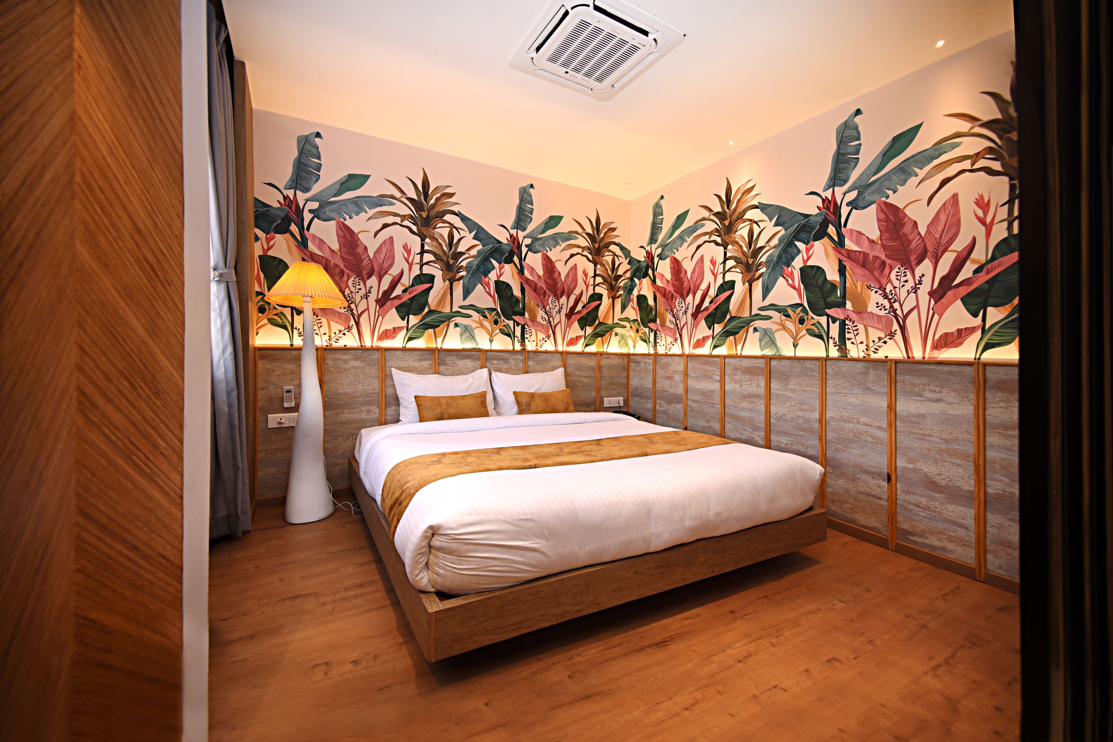 Une chambre avec un lit en bois, une table de chevet, une lampe et une fresque murale représentant des plantes tropicales.