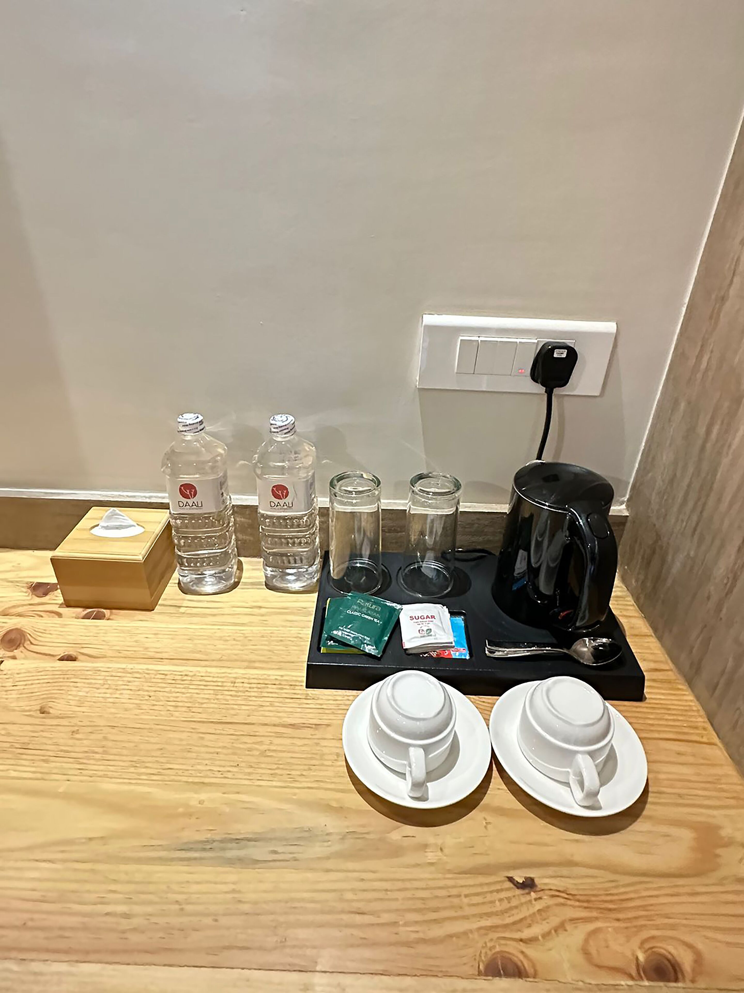 A wooden table with a coffee maker, two cups, two bottles of water, a tissue box, and a packet of sugar.