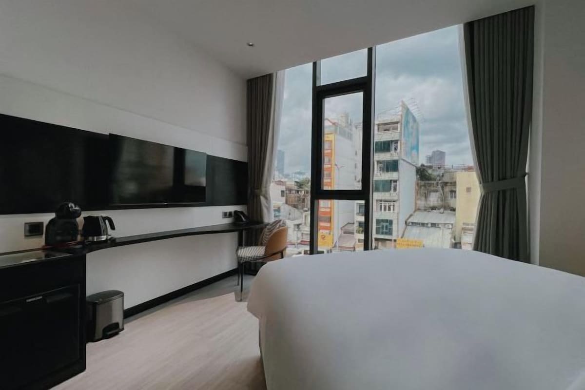 A modern hotel room with a large bed, a flat-screen TV, a desk with a coffee maker, and a view of the cityscape through large windows.