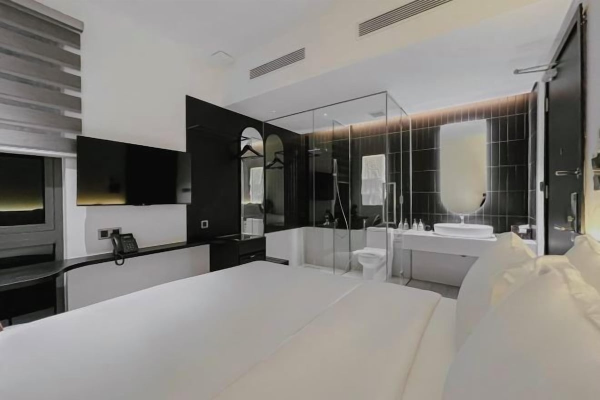 A modern hotel room with a large bed, a bedside table, a mirror, a TV, and a bathroom with a shower and toilet.