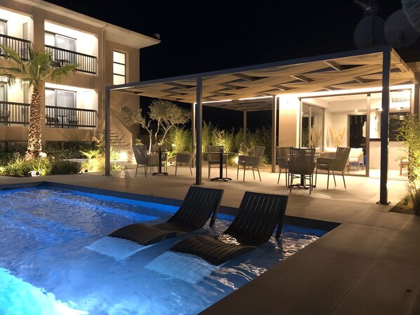A pool area with lounge chairs, a covered patio, and a building with balconies.