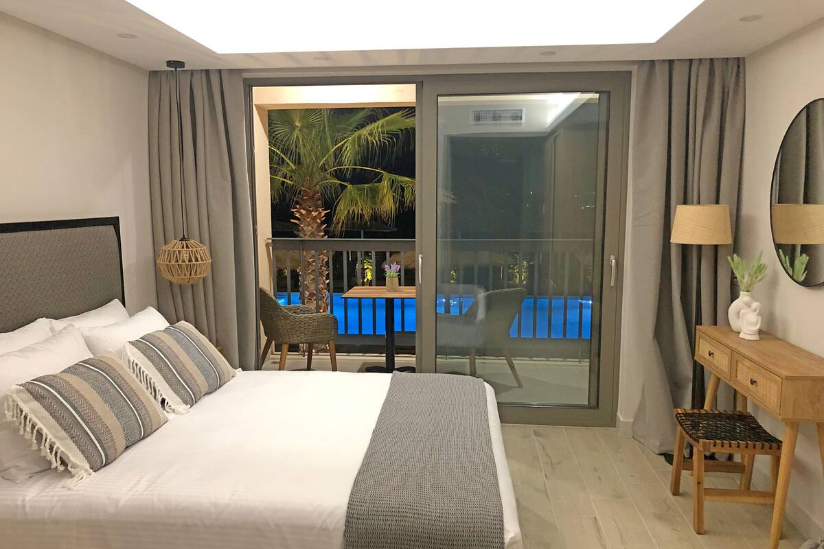 Ein Schlafzimmer mit einem großen Bett, einem Holznachttisch, einem Sessel und Blick auf einen Pool durch eine Glastür.