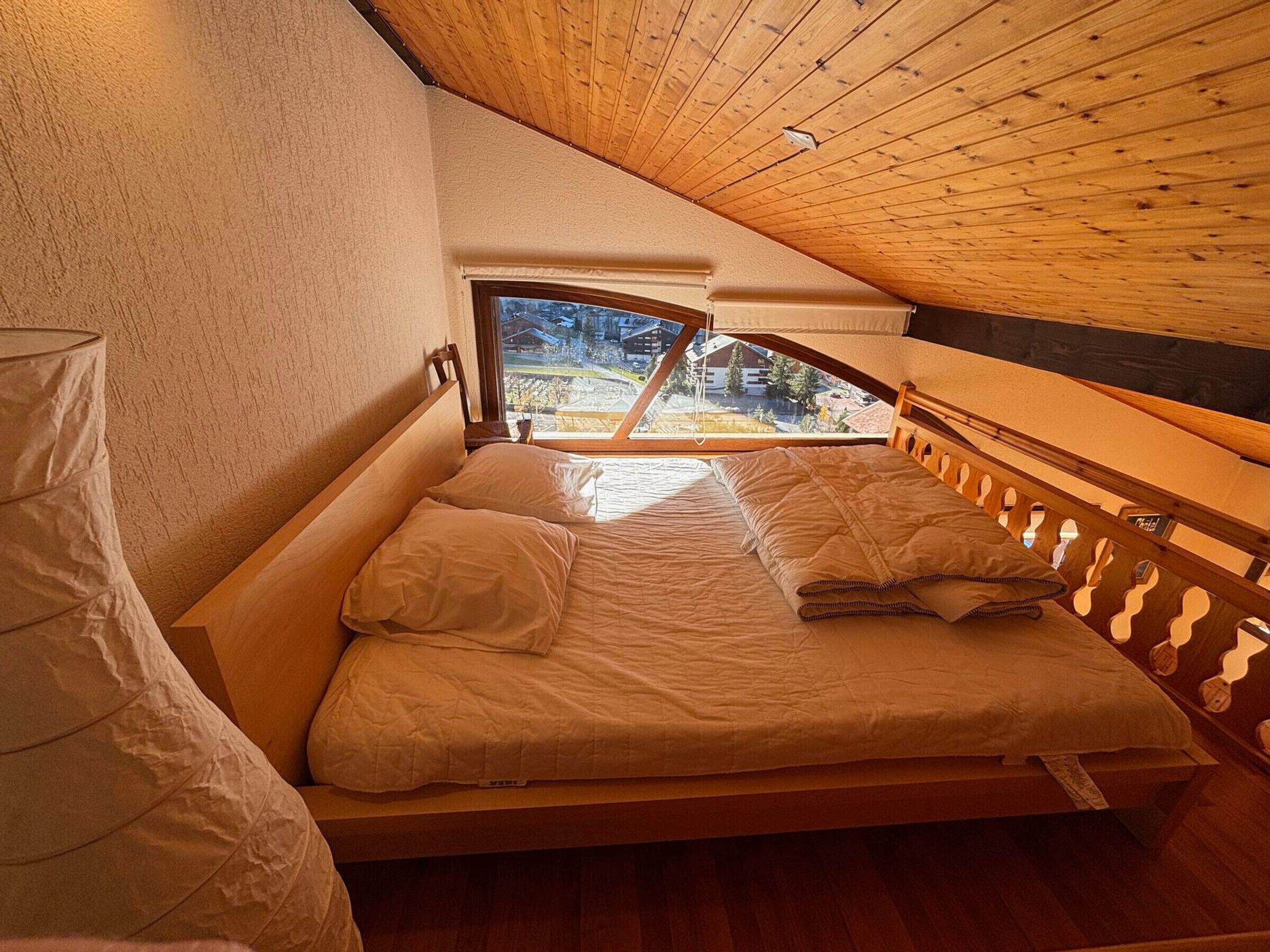 Een slaapkamer met een houten plafond, een bed met wit beddengoed en uitzicht op een woonwijk door een raam.