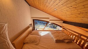 Une chambre avec un plafond en bois, un lit recouvert de draps blancs et une vue sur un quartier résidentiel par la fenêtre.