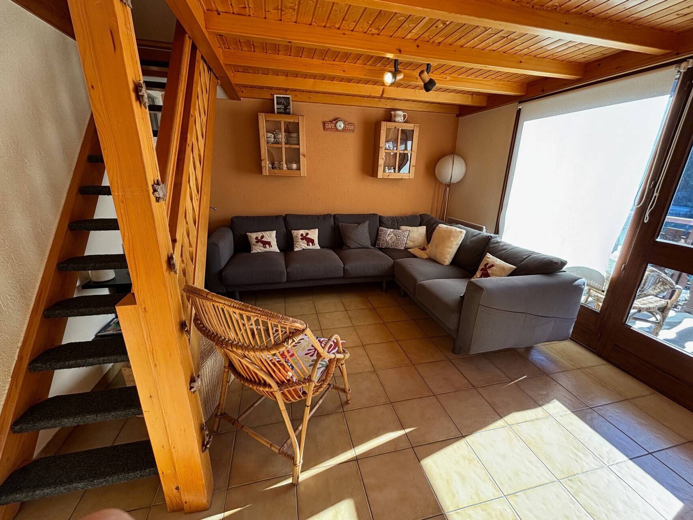 A cozy living room with a dark grey sectional sofa, a wicker chair, and a staircase with wooden railings.