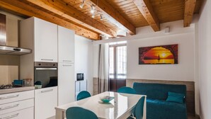 A modern kitchen with white cabinets, a built-in oven, and a dining area with a white table and teal chairs. A teal sofa is against the wall, and a painting of a sunset is visible. - Luna d Argento by Wonderful Italy - Nayra (Syracuse)