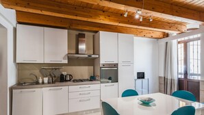 A modern kitchen with white cabinets, a stainless steel range hood, and a dining area with a white table and teal chairs. - Luna d Argento by Wonderful Italy - Nayra (Syracuse)