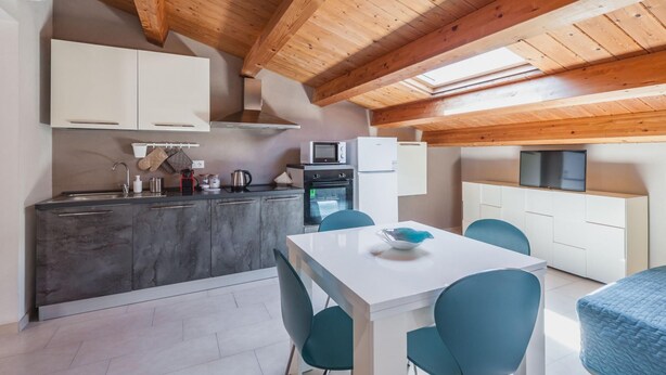 A modern kitchen with wooden beams, a dining area with teal chairs, and a TV mounted on the wall. - Luna d Argento by Wonderful Italy - Kimama (Syracuse)
