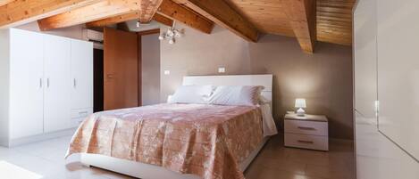 A bedroom with a wooden ceiling, a bed with a patterned bedspread, a bedside table with a lamp, and a wardrobe.