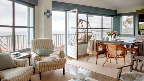 A cozy living room with striped armchairs, a coffee table with magazines, and a view of the ocean through large windows. - Wishome—A Fairytale Home to Extend Summer by the Sea (San Vicente de la Barquera)