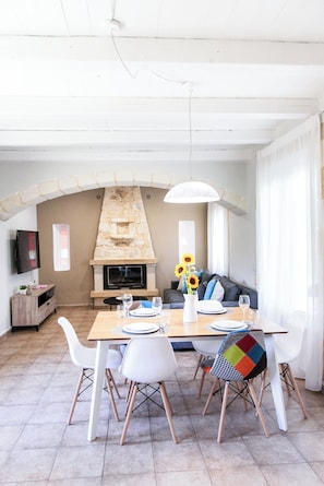 Un coin repas agrémenté d’une cheminée en pierre, de chaises blanches et d’une table dressée pour quatre personnes.