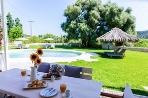 Une table avec des aliments pour le petit-déjeuner, un vase rempli de tournesols et une vue sur une piscine et des chaises longues.