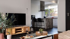 Un salon moderne doté d’un téléviseur à écran plat, d’une table basse avec des collations et des verres à vin, d’un meuble de rangement en bois et d’une grande fenêtre offrant une vue sur la cuisine et l’extérieur.