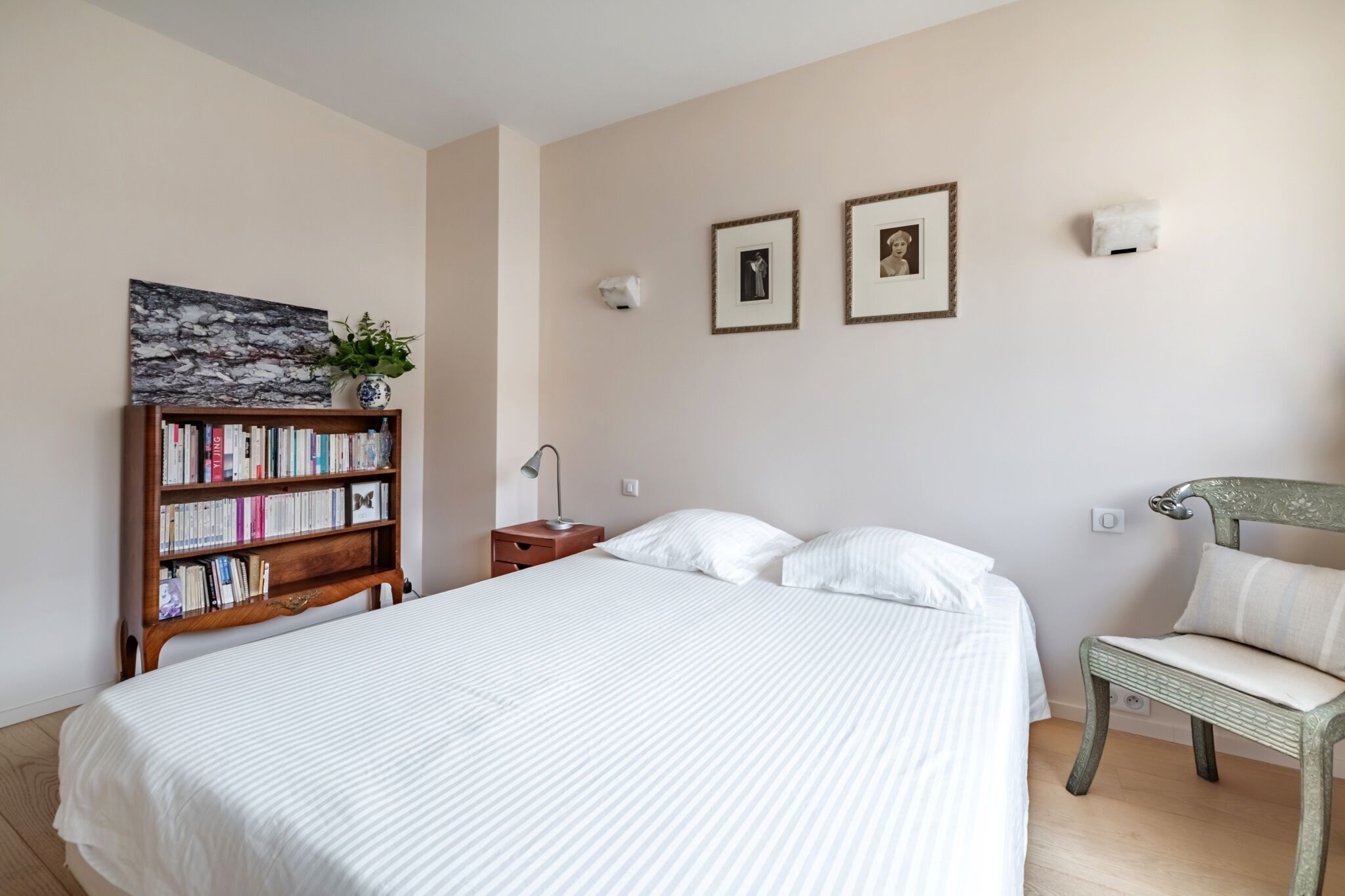 A neatly arranged bedroom with a large bed, a bookshelf, a chair, and two framed pictures on the wall.