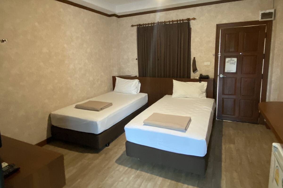 A hotel room with two single beds, a wooden floor, a dark brown door, and a window with curtains.