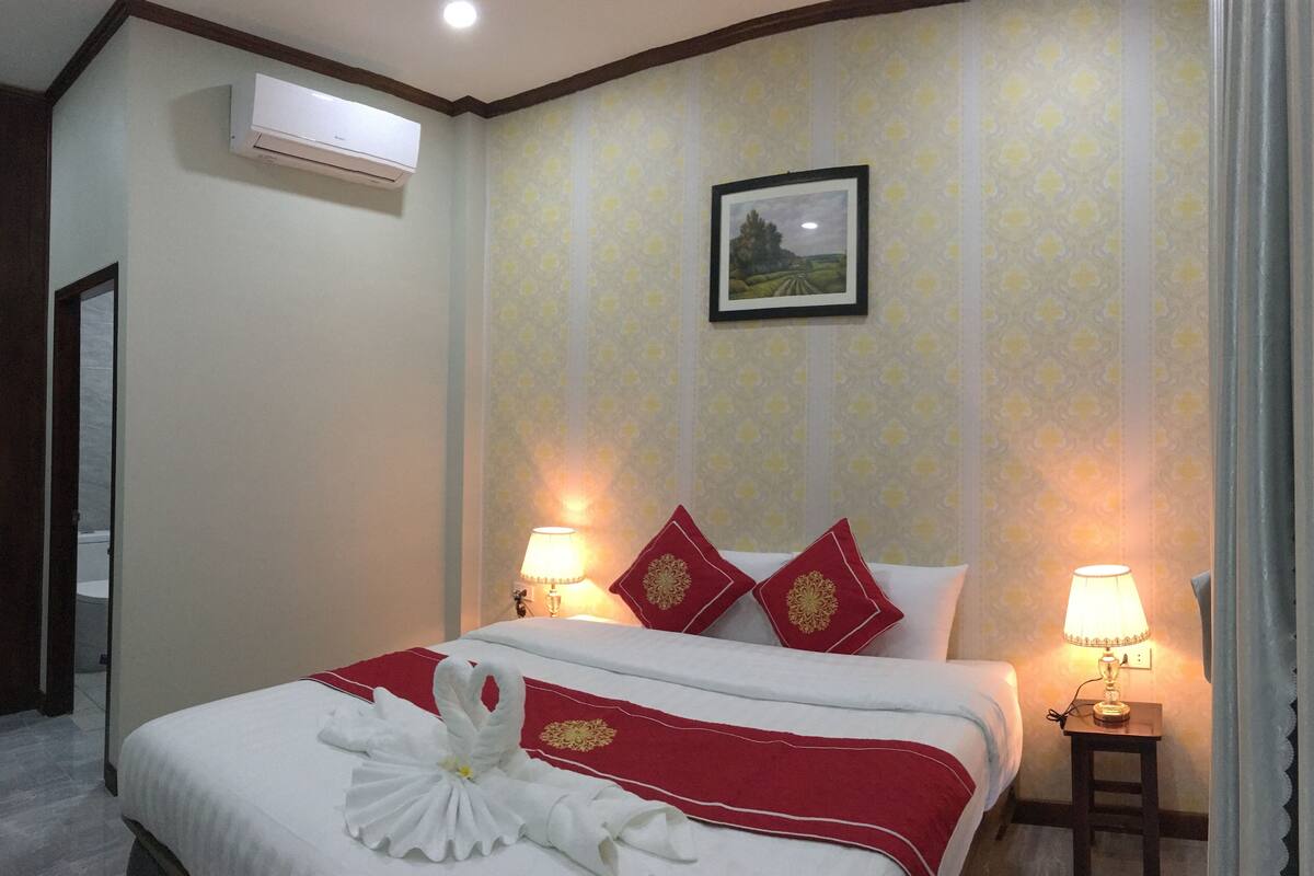 A hotel room with a bed, two bedside lamps, a framed picture on the wall, and an air conditioning unit.