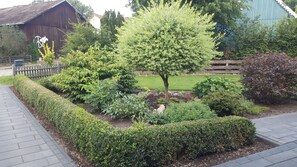 Een verzorgde tuin met diverse struiken en een centrale boom, begrensd door een houten omheining en een geplaveid pad.