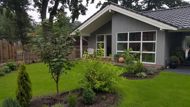 Une maison avec un toit en pente, de grandes fenêtres et un jardin bien entretenu, agrémenté de diverses plantes et d’une pelouse.