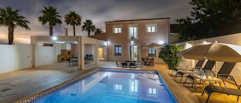 Una piscina con borde de azulejos, rodeada de sillones de descanso y sombrillas, con un edificio al fondo.