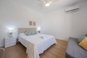 A bedroom with a bed, bedside table, lamp, air conditioning unit, and a sofa.
